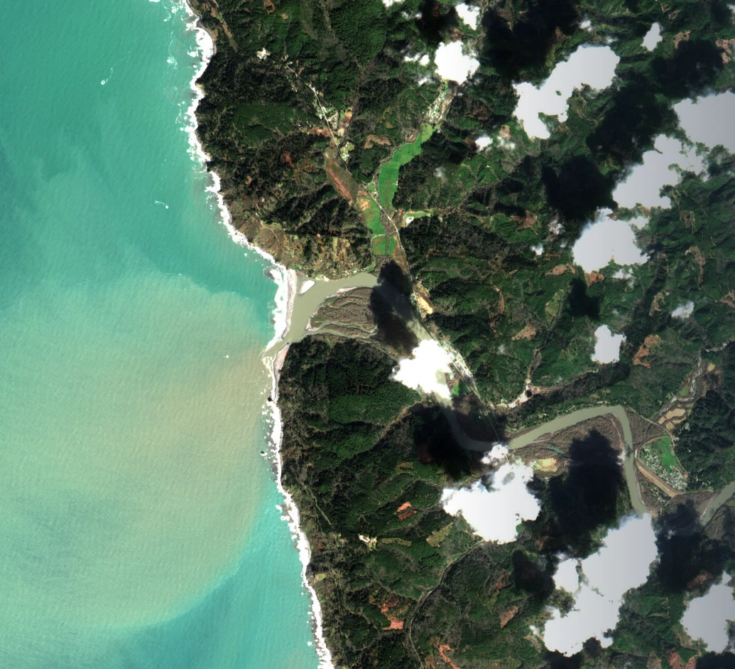 Satellite image of the Klamath River mouth: a pale sediment plume spreading into the turquoise Pacific beside forested hills and scattered clouds